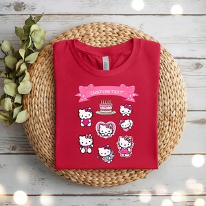 May include: A red t-shirt featuring multiple Hello Kitty designs, including a birthday cake and various poses. A pink banner with the text "CUSTOM TEXT" is above the designs. The shirt is displayed on a woven, circular placemat.