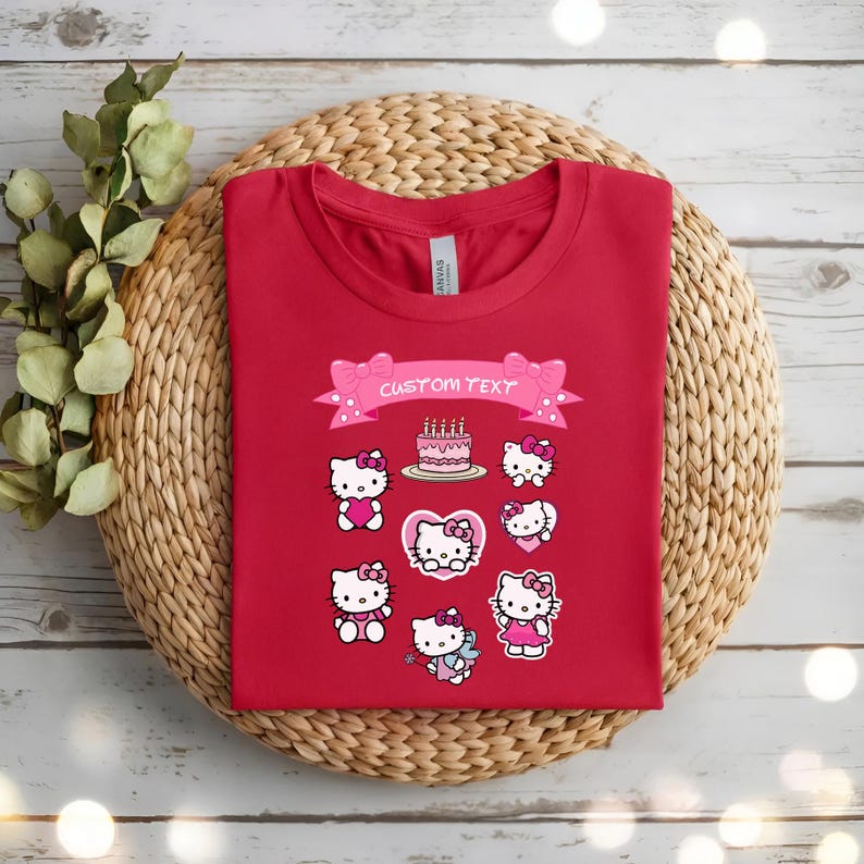 May include: A red t-shirt featuring multiple Hello Kitty designs, including a birthday cake and various poses. A pink banner with the text "CUSTOM TEXT" is above the designs. The shirt is displayed on a woven, circular placemat.
