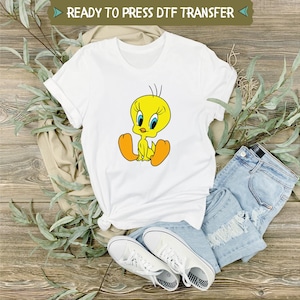 May include: White t-shirt with a yellow cartoon bird design. The bird is sitting with its feet crossed and has a blue beak and eyes. The shirt is ready to press with a DTF transfer.