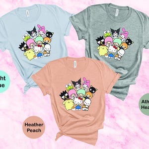 May include: Three t-shirts with a graphic print of a group of cartoon characters. The t-shirts are in light blue, heather peach, and athletic heather.
