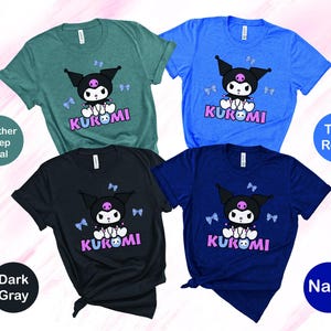 Puede incluir: Cuatro camisetas de manga corta en verde azulado, azul, gris oscuro y azul marino. Cada camiseta presenta un personaje de dibujos animados con cabeza negra, cuerpo blanco y la palabra "KUROMI" en rosa y morado. Las camisetas se muestran sobre un fondo blanco.
