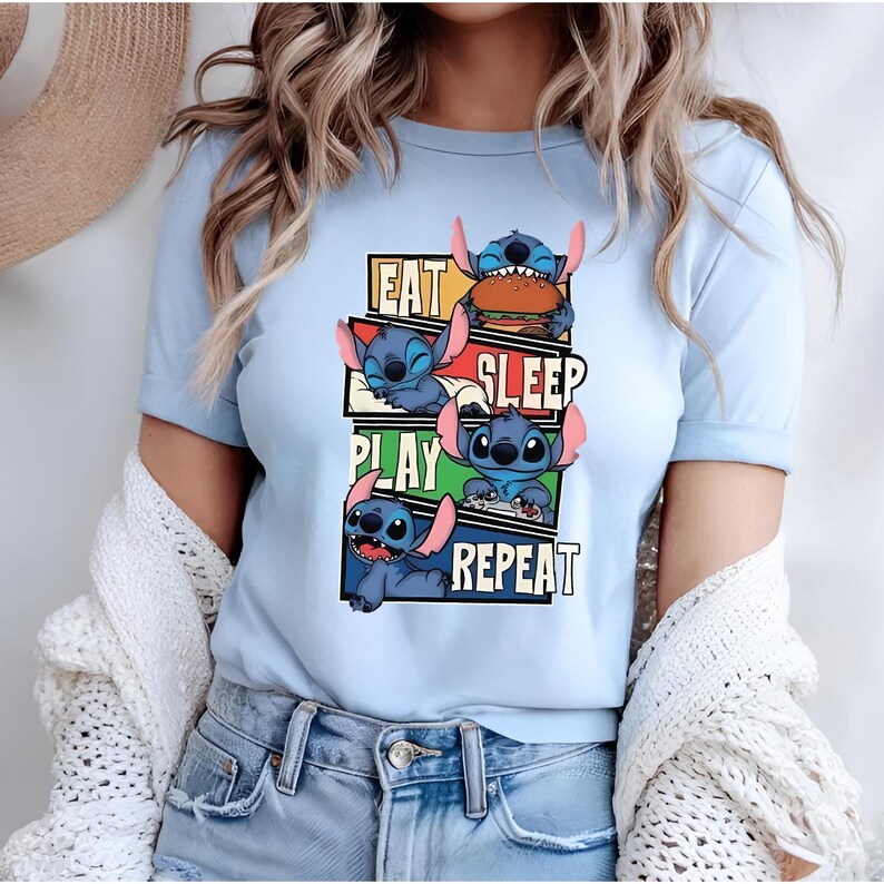 May include: Light blue t-shirt featuring the character Stitch from Lilo & Stitch. The shirt has a graphic design with the words "EAT SLEEP PLAY REPEAT" and images of Stitch in various poses. The shirt is short-sleeved.
