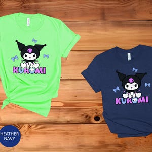 Puede incluir: Dos camisetas de manga corta, una verde menta y otra azul marino, con un personaje de dibujos animados con orejas negras, una calavera rosa y la palabra "KUROMI" en rosa y azul. El personaje est&aacute; rodeado de lazos azules.