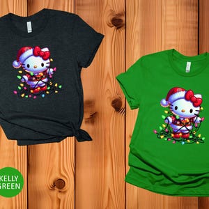 May include: Two short-sleeved t-shirts, one heather dark grey and one kelly green, feature a festive graphic. The graphic is of a cartoon cat wearing a Santa hat and wrapped in Christmas lights. The cat has a red bow and is surrounded by colorful lights.
