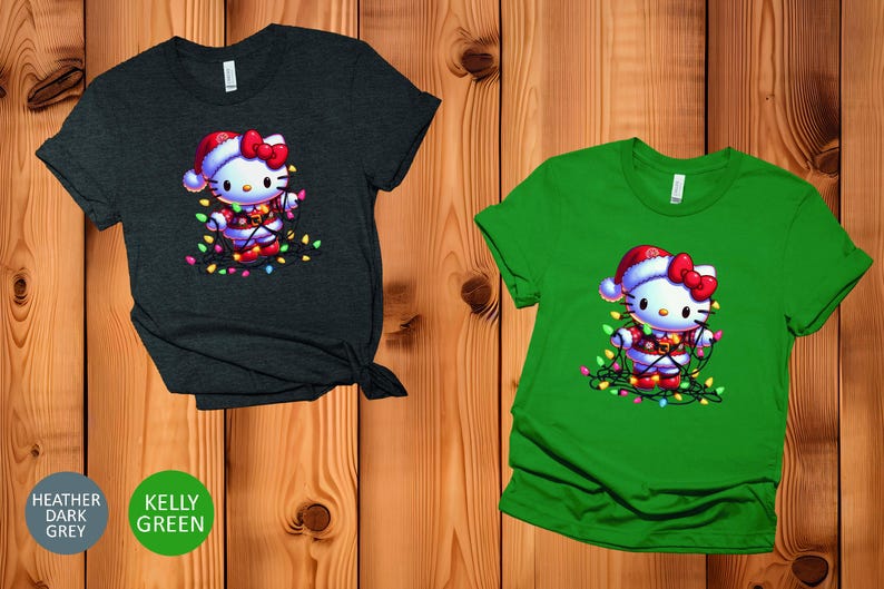 May include: Two short-sleeved t-shirts, one heather dark grey and one kelly green, feature a festive graphic. The graphic is of a cartoon cat wearing a Santa hat and wrapped in Christmas lights. The cat has a red bow and is surrounded by colorful lights.