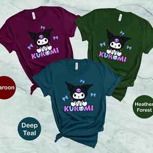 Puede incluir: Tres camisetas de manga corta en color burdeos, verde azulado oscuro y verde bosque jaspeado. Cada camiseta presenta un gr&aacute;fico de un personaje negro con una calavera rosa, lazos azules y la palabra "KUROMI" en rosa y morado.