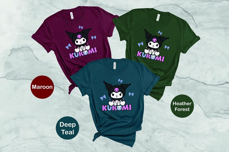Puede incluir: Tres camisetas de manga corta en color burdeos, verde azulado oscuro y verde bosque jaspeado. Cada camiseta presenta un gr&aacute;fico de un personaje negro con una calavera rosa, lazos azules y la palabra "KUROMI" en rosa y morado.