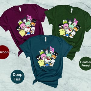 May include: Three t-shirts with a graphic print of cartoon characters. The t-shirts are in maroon, deep teal, and heather forest colors. The graphic features a group of cartoon characters with a pink, green, and blue color scheme.