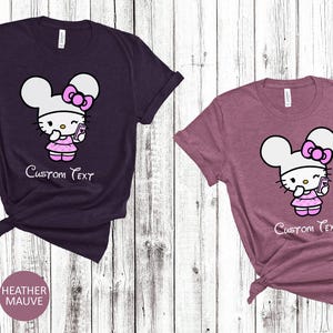Pu&ograve; includere: Due magliette a maniche corte, una color melange marrone e una color melange malva, ciascuna con un personaggio dei cartoni animati. Il personaggio ha una testa bianca, un fiocco rosa e un vestito, che tiene in mano un telefono. Il testo "Custom Text" &egrave; sotto.