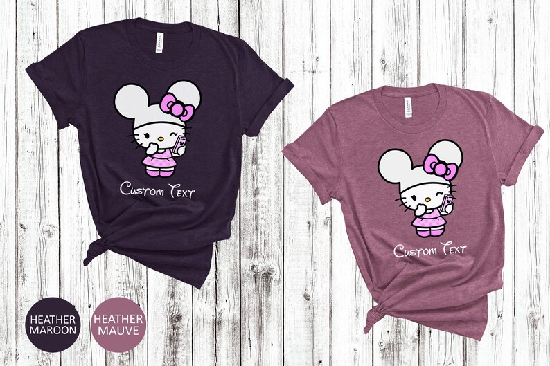 Pu&ograve; includere: Due magliette a maniche corte, una color melange marrone e una color melange malva, ciascuna con un personaggio dei cartoni animati. Il personaggio ha una testa bianca, un fiocco rosa e un vestito, che tiene in mano un telefono. Il testo "Custom Text" &egrave; sotto.