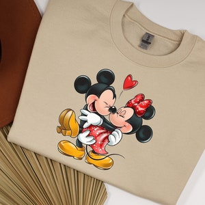 May include: A beige sweatshirt with a cartoon image of Mickey Mouse and Minnie Mouse hugging. Mickey is wearing red shorts and yellow shoes. Minnie is wearing a red dress with white polka dots and yellow shoes. A red heart is floating above their heads.