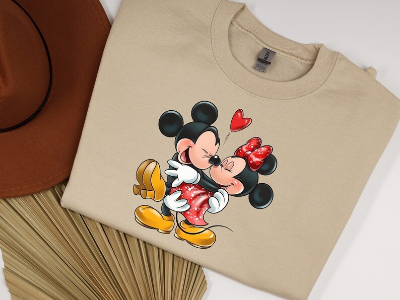 May include: A beige sweatshirt with a cartoon image of Mickey Mouse and Minnie Mouse hugging. Mickey is wearing red shorts and yellow shoes. Minnie is wearing a red dress with white polka dots and yellow shoes. A red heart is floating above their heads.