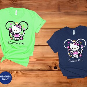 May include: Two t-shirts with a graphic of Hello Kitty dressed as a princess with a wand and a crown. The graphic is inside a Mickey Mouse ear shape. The t-shirts are in mint green and navy blue. The text "Custom Text" is below the graphic.
