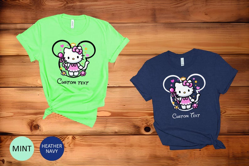 May include: Two t-shirts with a graphic of Hello Kitty dressed as a princess with a wand and a crown. The graphic is inside a Mickey Mouse ear shape. The t-shirts are in mint green and navy blue. The text "Custom Text" is below the graphic.
