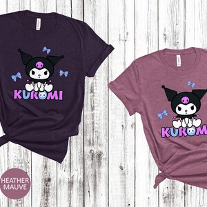 Puede incluir: Dos camisetas de manga corta, una granate jaspeado y otra malva jaspeado, cada una con un personaje de dibujos animados. El personaje tiene una cabeza negra, un cuerpo blanco y la palabra "KUROMI" en morado. Hay lazos azul claro dispersos.