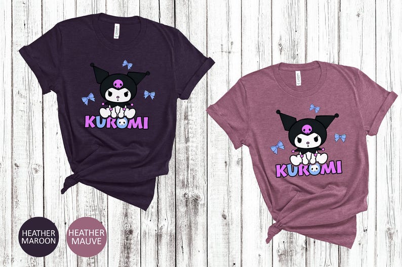 Puede incluir: Dos camisetas de manga corta, una granate jaspeado y otra malva jaspeado, cada una con un personaje de dibujos animados. El personaje tiene una cabeza negra, un cuerpo blanco y la palabra "KUROMI" en morado. Hay lazos azul claro dispersos.