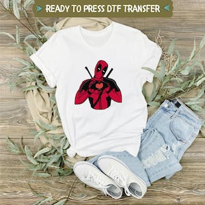 May include: White t-shirt featuring a graphic of Deadpool with a heart-shaped design. The character is depicted in red and black, holding a heart. The text "READY TO PRESS DTF TRANSFER" is at the top. The shirt is paired with blue jeans and white sneakers.