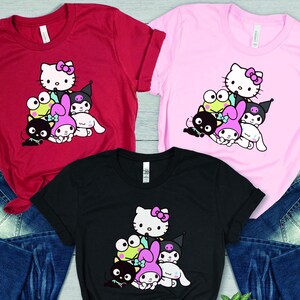K&ouml;nnte beinhalten: Drei T-Shirts mit einem Grafikdruck von Hello Kitty und ihren Freunden. Die Shirts sind in Rot, Pink und Schwarz erh&auml;ltlich.