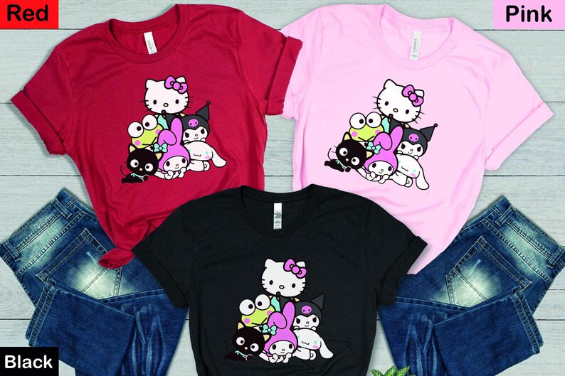 K&ouml;nnte beinhalten: Drei T-Shirts mit einem Grafikdruck von Hello Kitty und ihren Freunden. Die Shirts sind in Rot, Pink und Schwarz erh&auml;ltlich.