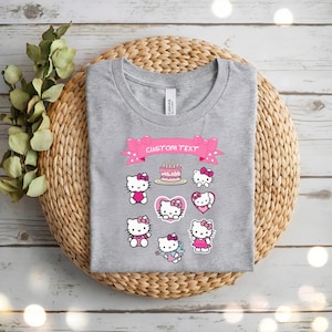 May include: A gray t-shirt featuring multiple Hello Kitty designs with pink accents. The shirt includes a pink banner with the text "CUSTOM TEXT" and a birthday cake graphic. The shirt is displayed on a woven basket.