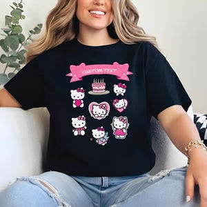 May include: Black t-shirt featuring a pink banner with the text "CUSTOM TEXT" and pink bows. The shirt has multiple illustrations of a cartoon cat with a pink bow, hearts, and a birthday cake.