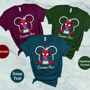 May include: Three short-sleeved t-shirts in maroon, deep teal, and heather forest green. Each shirt features a cartoon Spider-Man character with a birthday cake and the text "Custom Text" in white. The Spider-Man design is set within a white Mickey Mouse-shaped outline.