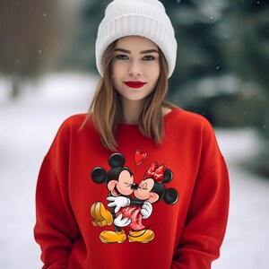 May include: A red sweatshirt with a graphic print of Mickey Mouse and Minnie Mouse kissing. The sweatshirt has a crew neck and long sleeves.
