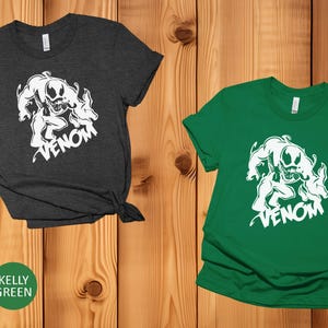 May include: Two t-shirts with a white graphic of Venom, a Marvel Comics character. The t-shirt on the left is heather dark grey and the t-shirt on the right is kelly green.
