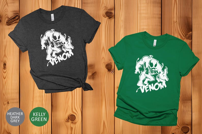 May include: Two t-shirts with a white graphic of Venom, a Marvel Comics character. The t-shirt on the left is heather dark grey and the t-shirt on the right is kelly green.