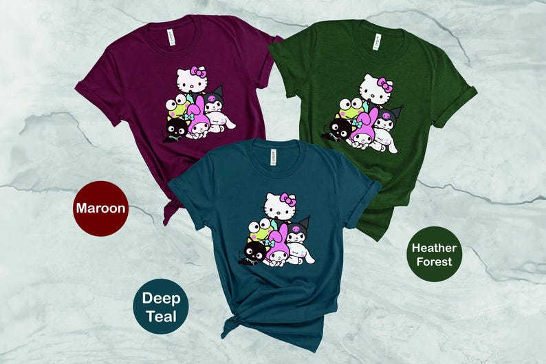 K&ouml;nnte beinhalten: Drei T-Shirts mit unterschiedlichen Hintergrundfarben und einem grafischen Druck einer Gruppe von Comicfiguren. Zu den Figuren geh&ouml;ren ein rosa-wei&szlig;es Hello-Kitty-&auml;hnliches Charakter, ein gr&uuml;ner Frosch, eine schwarz-wei&szlig;e Katze und ein rosa Kaninchen. Die T-Shirts sind in den Farben Rotbraun, Dunkelpetrol und Heidekrautwald.