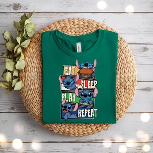 May include: A green t-shirt featuring the character Stitch from Lilo & Stitch. The shirt has the words "EAT SLEEP PLAY REPEAT" with Stitch in various poses. The shirt is displayed on a woven mat.