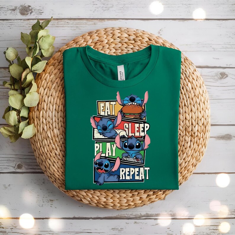 May include: A green t-shirt featuring the character Stitch from Lilo & Stitch. The shirt has the words "EAT SLEEP PLAY REPEAT" with Stitch in various poses. The shirt is displayed on a woven mat.