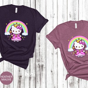 May include: Two short-sleeved t-shirts, one heather maroon and one heather mauve, featuring a cartoon princess cat design. The cat wears a crown and dress, with a rainbow and stars in the background. The shirts are knotted at the bottom.