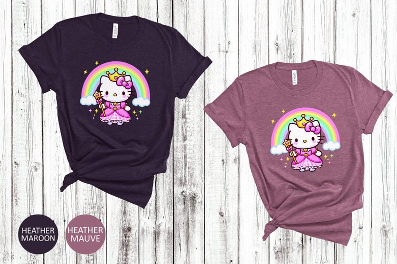 May include: Two short-sleeved t-shirts, one heather maroon and one heather mauve, featuring a cartoon princess cat design. The cat wears a crown and dress, with a rainbow and stars in the background. The shirts are knotted at the bottom.