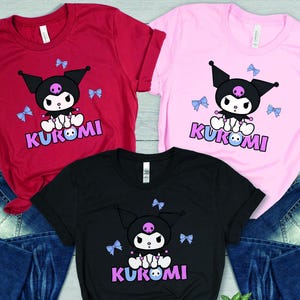 Puede incluir: Tres camisetas en rojo, rosa y negro, cada una con un personaje de dibujos animados con cabeza negra, una calavera y la palabra "KUROMI" en morado. Lazos azul claro est&aacute;n dispersos alrededor del personaje. Las camisetas se combinan con vaqueros azules.