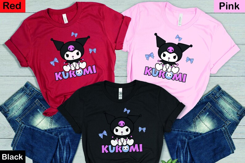 Puede incluir: Tres camisetas en rojo, rosa y negro, cada una con un personaje de dibujos animados con cabeza negra, una calavera y la palabra "KUROMI" en morado. Lazos azul claro est&aacute;n dispersos alrededor del personaje. Las camisetas se combinan con vaqueros azules.