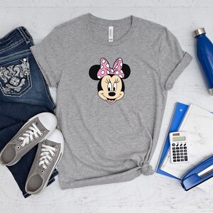 May include: A gray t-shirt with a cartoon image of Minnie Mouse wearing a pink bow with white polka dots. The shirt is laid out on a white wooden surface with a pair of gray sneakers, blue jeans, a blue water bottle, a blue notebook, a calculator, and a blue stapler.