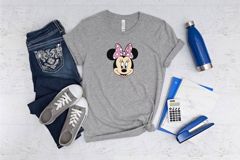 May include: A gray t-shirt with a cartoon image of Minnie Mouse wearing a pink bow with white polka dots. The shirt is laid out on a white wooden surface with a pair of gray sneakers, blue jeans, a blue water bottle, a blue notebook, a calculator, and a blue stapler.