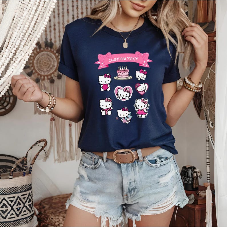 May include: Navy blue t-shirt featuring a collection of Hello Kitty graphics. The design includes various poses of the character, a birthday cake, and a banner with the text "CUSTOM TEXT". The shirt has short sleeves and a classic crew neck.