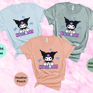 Puede incluir: Tres camisetas de manga corta en azul claro, melocot&oacute;n jaspeado y gris deportivo jaspeado. Cada camiseta presenta un personaje de dibujos animados con orejas negras, una calavera y la palabra "KUROMI" en rosa y morado.