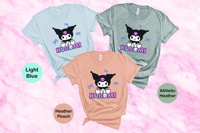 Puede incluir: Tres camisetas de manga corta en azul claro, melocot&oacute;n jaspeado y gris deportivo jaspeado. Cada camiseta presenta un personaje de dibujos animados con orejas negras, una calavera y la palabra "KUROMI" en rosa y morado.