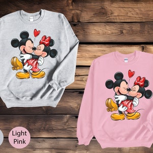 May include: A gray sweatshirt with a graphic of Mickey Mouse and Minnie Mouse hugging. The sweatshirt is available in ash and light pink.