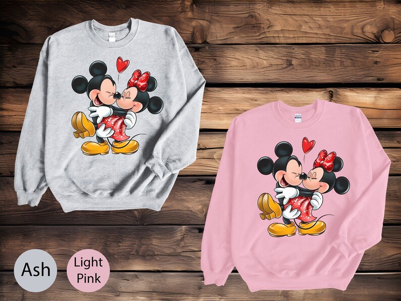 May include: A gray sweatshirt with a graphic of Mickey Mouse and Minnie Mouse hugging. The sweatshirt is available in ash and light pink.