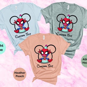 May include: Three t-shirts in light blue, athletic heather, and heather peach. Each shirt features a cartoon Spider-Man character with a birthday cake and the text "Custom Text". The design is framed by a Mickey Mouse-style outline.