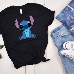 May include: Black t-shirt with a blue Stitch character from Lilo & Stitch. The character is sitting with its arms crossed and has a light blue body and purple ears.
