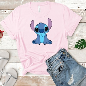 May include: A pink t-shirt with a blue cartoon character of Stitch from Lilo & Stitch sitting on the front of the shirt. The shirt is on a wooden surface with a pair of silver sandals, a pair of red sunglasses, and a green leaf keychain.