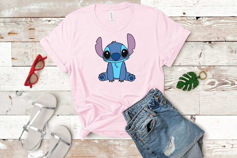 May include: A pink t-shirt with a blue cartoon character of Stitch from Lilo & Stitch sitting on the front of the shirt. The shirt is on a wooden surface with a pair of silver sandals, a pair of red sunglasses, and a green leaf keychain.