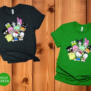 May include: Two t-shirts with a graphic print of a group of cartoon characters. The shirt on the left is heather dark grey and the shirt on the right is kelly green.