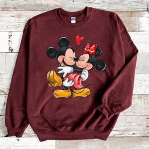 May include: A maroon sweatshirt with a graphic print of Mickey Mouse and Minnie Mouse hugging. Mickey is wearing yellow shoes and Minnie is wearing red shoes with white polka dots. A red heart balloon is above them.