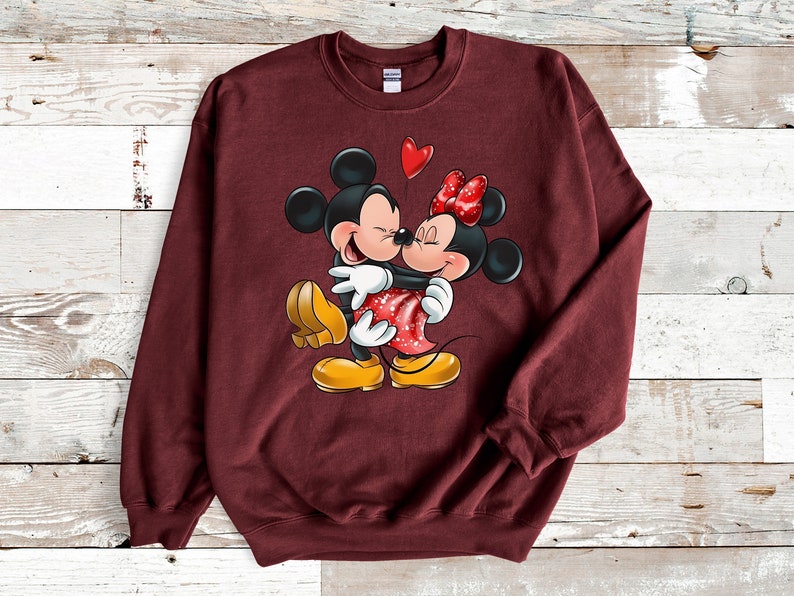 May include: A maroon sweatshirt with a graphic print of Mickey Mouse and Minnie Mouse hugging. Mickey is wearing yellow shoes and Minnie is wearing red shoes with white polka dots. A red heart balloon is above them.